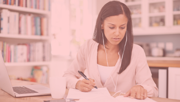 Woman writing in a notebook, with wired earpods, in an office with bookcases and an open laptop.