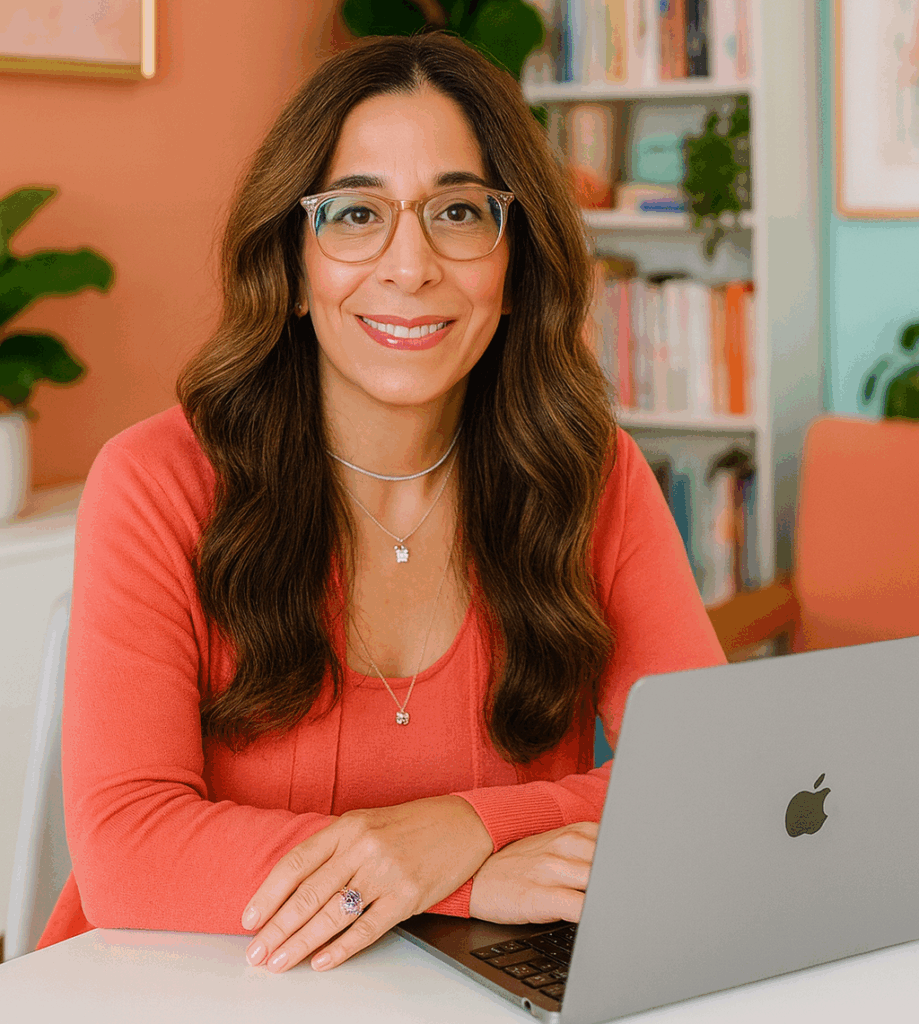 Natalie Anderson sits at a white desk with a MacBook Pro, wearing a coral long-sleeved top and silver necklaces, including one with a small lucky cat charm. She has a pink manicure and an amethyst ring on her left hand, smiling in a bright room with coral and teal walls, bookshelves, art, and plants.