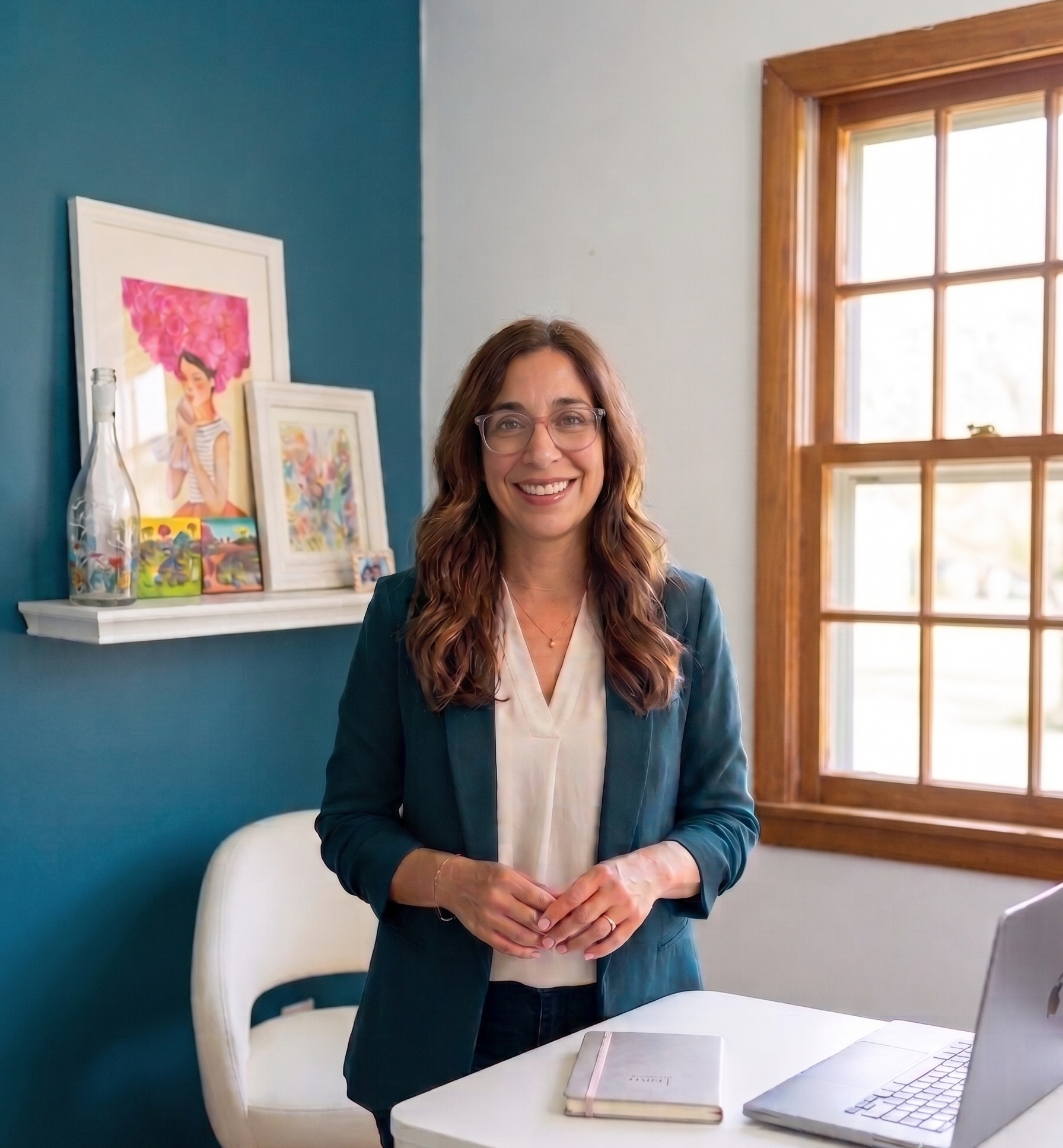 Natalie Anderson standing at her desk in her home office, wearing a teal blazer, with a laptop and notebook in front of her